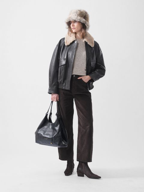 Una donna con un cappello di pelliccia, una giacca di pelle nera, un maglione beige, pantaloni marroni e stivali con tacco, che tiene una grande borsa nera e sta in piedi davanti a uno sfondo bianco.