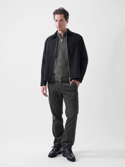 Un uomo si trova di fronte a uno sfondo a tinta unita, indossa una giacca nera, un maglione grigio con cerniera, pantaloni scuri e scarpe nere, e guarda leggermente di lato.