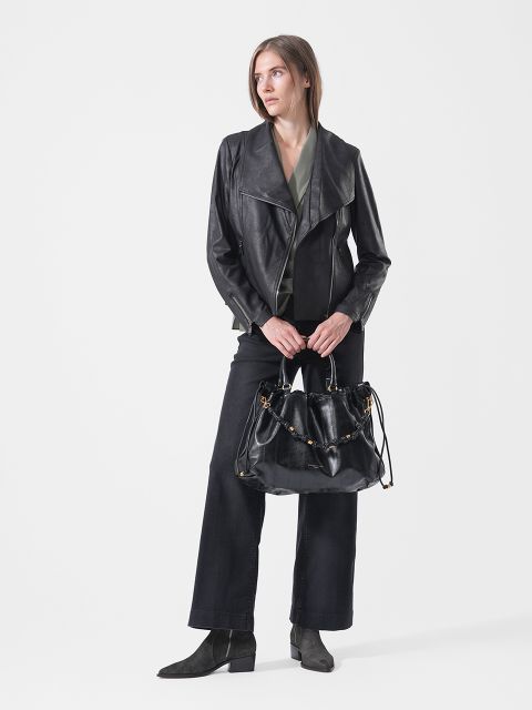 Una donna con una giacca di pelle nera, pantaloni neri e stivali è in piedi con una grande borsa nera in mano davanti a uno sfondo bianco, guardando di lato.