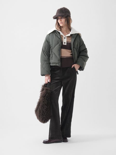 Una donna con una giacca a vento verde, un maglione a righe, pantaloni marroni e un cappello di pelliccia, che tiene in mano una soffice borsa marrone e sta in piedi davanti a uno sfondo bianco semplice.