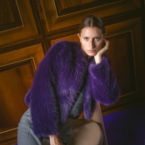 Una persona con un cappotto di pelliccia sintetica viola è appoggiata a una sedia, guarda la telecamera, con pareti rivestite in legno sullo sfondo.