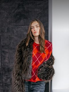 Una donna indossa un maglione argyle rosso-arancione sotto una giacca scura di pelliccia sintetica.