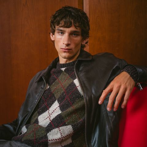 Un giovane uomo con una giacca di pelle scura e un maglione argyle è seduto su una sedia rossa, in posa davanti a una parete di legno e guarda la telecamera.