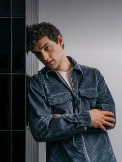 Un uomo che indossa una camicia di velluto a coste blu con cerniera, con le braccia incrociate e appoggiato a una parete di piastrelle scure.