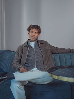 Un uomo seduto su un divano, che indossa una giacca grigia con cerniera, una camicia bianca e pantaloni bianchi.