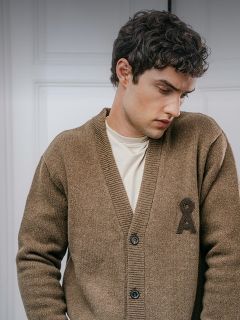 Un uomo indossa un cardigan marrone con bottoni e un emblema scuro ricamato sul petto sopra una maglietta color crema.