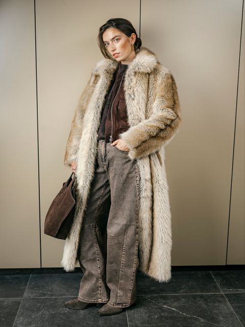 Una donna con un lungo cappotto di pelliccia, pantaloni marroni e stivali è in piedi in una stanza, tiene in mano una borsa marrone e guarda la telecamera con un'espressione impassibile.