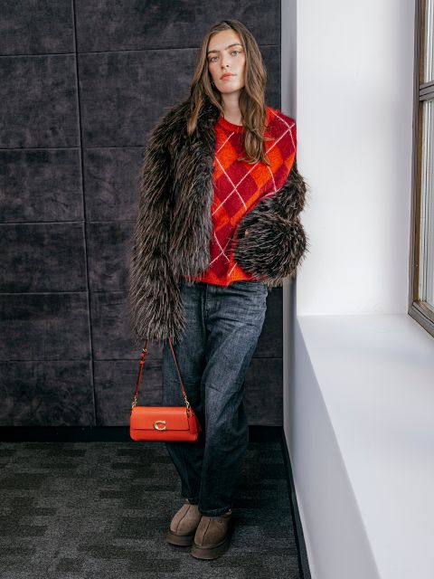 Una donna con un maglione rosso a rombi, un cappotto di pelliccia sintetica, jeans grigi e scarpe marrone chiaro è appoggiata a una parete vicino a una finestra e tiene in mano una borsa rossa.