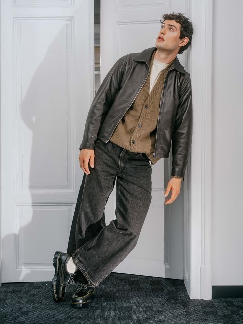 Un uomo con pantaloni scuri, una giacca marrone e un cappotto di pelle è appoggiato a una porta bianca e guarda in alto con un'espressione sorpresa.