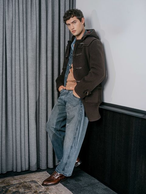 Un giovane uomo con un cappotto marrone, un maglione arancione e jeans blu è appoggiato a un muro con le mani in tasca, in piedi accanto a tende grigie.