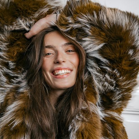 Donna sorridente con un cappotto di pelliccia sintetica marrone e bianca, con lunghi capelli sciolti, che posa con una mano sopra la testa davanti a uno sfondo chiaro.