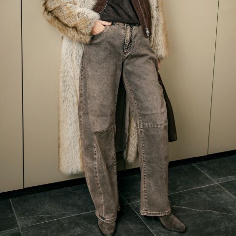 Una persona che indossa jeans marroni a gamba larga, un cappotto di pelliccia e stivali a punta, con una borsa in mano, è in piedi davanti a degli armadietti beige.