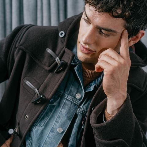 Un uomo con un cappotto scuro e una giacca di jeans si tocca la tempia e guarda pensieroso in basso davanti a tende grigie.