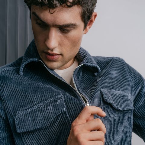 Un uomo con riccioli corti, che indossa una giacca di velluto a coste blu sopra una camicia bianca, guarda in basso mentre chiude la cerniera della giacca.