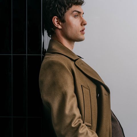 Un uomo con un cappotto marrone è in piedi di profilo davanti a un muro in bianco e nero, con un'espressione seria, e guarda avanti.