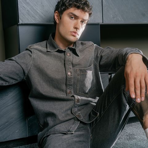Un uomo con riccioli corti, che indossa una camicia di jeans grigia e jeans, è seduto sul pavimento, appoggiato a una parete scura e moderna, e guarda la fotocamera.
