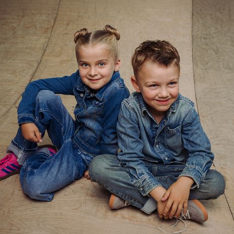 Due bambini in abiti di jeans sono seduti su un pavimento di legno e sorridono alla telecamera. Uno indossa scarpe da ginnastica rosa, l'altro scarpe marrone chiaro.