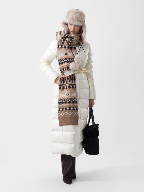 Una donna indossa un lungo piumino bianco, una sciarpa marrone fantasia con il logo UGG e un cappello da trapper in pelliccia sintetica; in mano tiene una borsa nera.