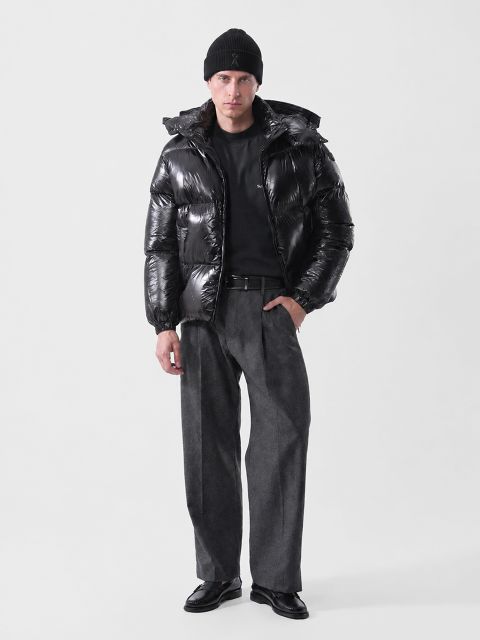 Un uomo che indossa un berretto nero, un piumino nero lucido, una camicia nera, pantaloni grigi e mocassini neri.