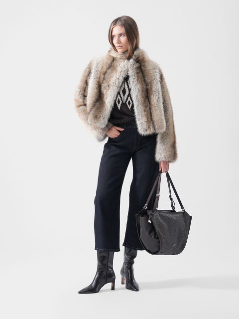 Una modella indossa una giacca di pelliccia sopra un maglione a fantasia, jeans scuri a gamba larga e stivali neri con tacco, e porta una grande borsa a tracolla nera.