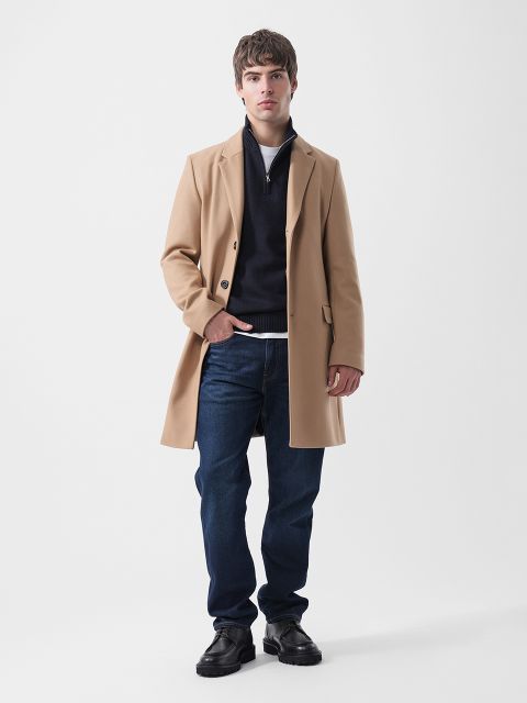 Un uomo che indossa un cappotto marrone chiaro, un maglione blu navy con cerniera sul colletto, una maglietta bianca, jeans blu scuro e scarpe nere.