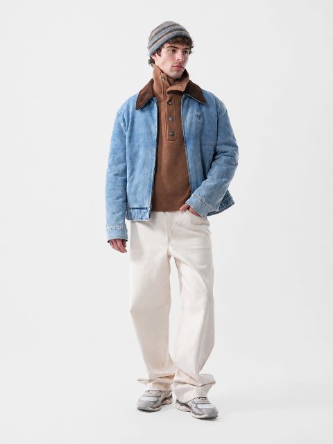 Un uomo che indossa una giacca di jeans blu sopra un maglione marrone con bottoni, pantaloni color crema e un berretto.