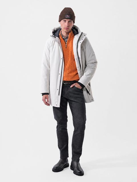Un uomo che indossa un parka bianco con cappuccio, un maglione arancione con cerniera a un quarto, jeans neri e un cappello marrone.