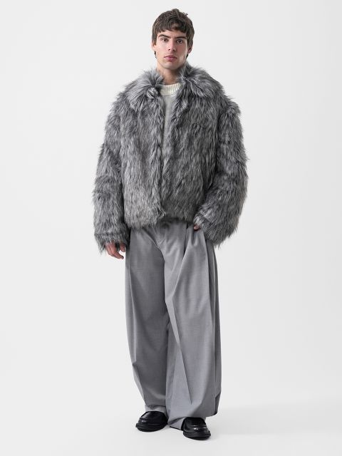 Un uomo che indossa una giacca di pelliccia sintetica grigia, un maglione color crema e pantaloni grigi a gamba larga.