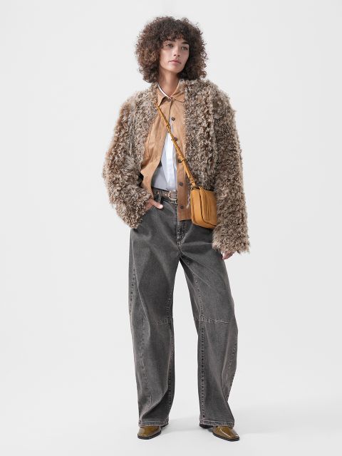 Una persona che indossa una giacca dalla texture soffice sopra una camicia marrone chiaro con colletto bianco, abbinata a jeans grigi a gamba larga e una borsa a tracolla marrone chiaro.