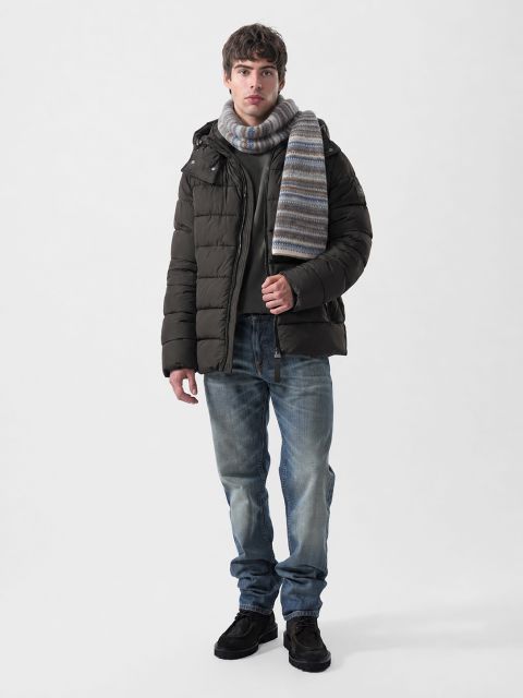Un uomo che indossa una giacca piumino marrone scuro, un maglione grigio, una sciarpa a righe, jeans blu e scarpe scure.