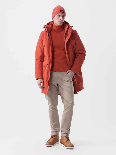 Un uomo che indossa un berretto arancione, un dolcevita e un parka, oltre a pantaloni beige e stivali di pelle marroni.