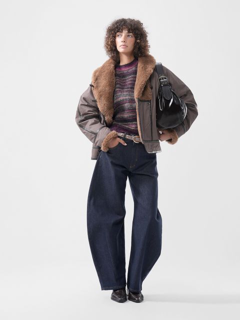 Una persona che indossa una giacca marrone con colletto di pelliccia, un maglione a righe viola e jeans scuri a gamba larga, con una borsa a tracolla nera.