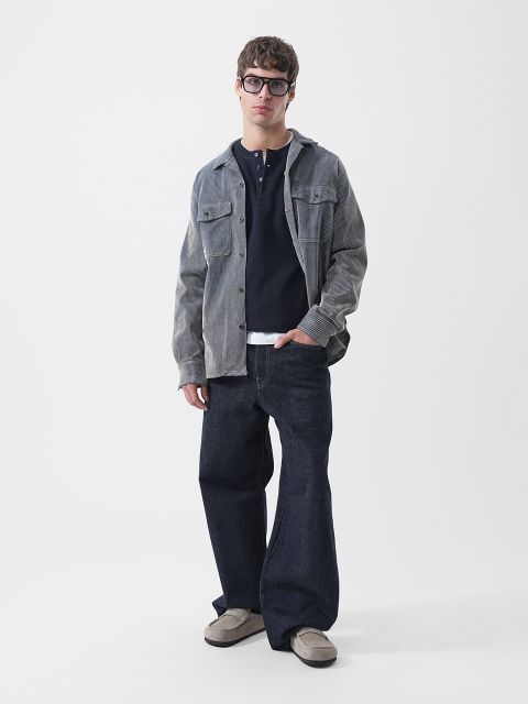 Un uomo con occhiali dalla montatura spessa, una camicia di velluto a coste sbottonata sopra una maglietta Henley blu navy, jeans larghi blu scuro e mocassini grigi.