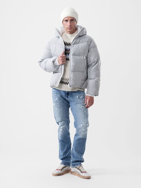 Un uomo che indossa una giacca piumino grigio chiaro, un maglione fantasia, un berretto bianco e jeans blu slavato, oltre a scarpe da ginnastica.