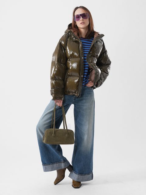 Una persona che indossa un piumino lucido verde oliva, una camicia a righe blu, jeans a gamba larga, occhiali da sole viola e tiene in mano una borsa verde oliva.