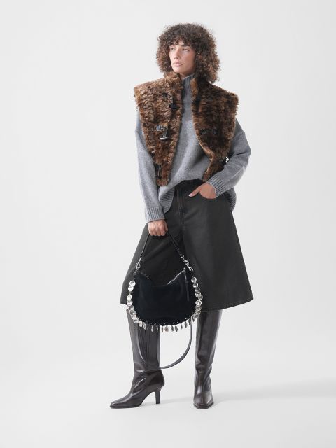 Una modella indossa un dolcevita grigio, un gilet di pelliccia sintetica marrone e una gonna di jeans nera; tiene anche una borsa nera con dettagli rotondi argentati e indossa stivali alti marroni.