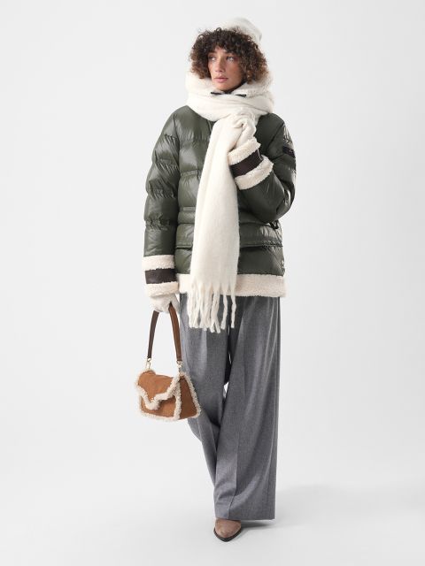 Una persona che indossa un piumino verde oliva con finiture in pelle di agnello, una sciarpa e un cappello bianchi, pantaloni grigi e porta una piccola borsa marrone con finiture in pelle di agnello.