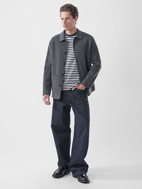 Un uomo che indossa una giacca grigia sopra una maglietta a righe blu navy e bianche, jeans blu scuro e mocassini neri.
